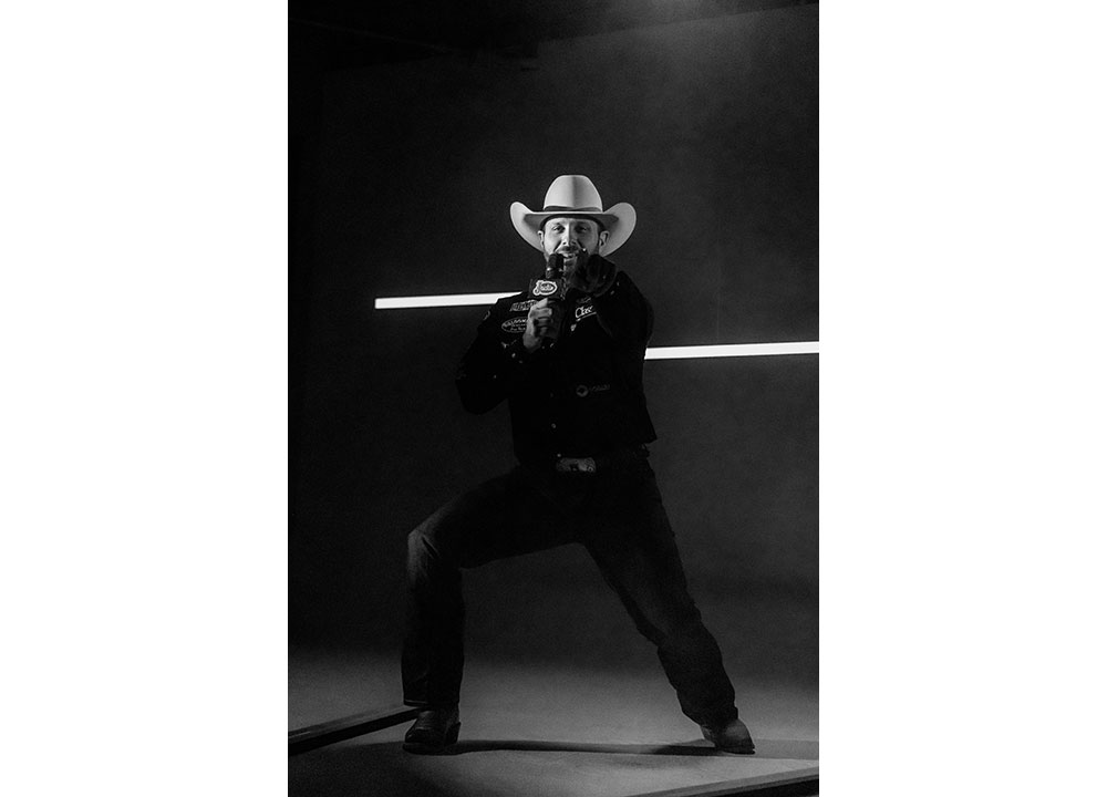 Anthony Lucia vistiendo una camisa negra y un sombrero de vaquero, sosteniendo un micrófono y posando mientras apunta a la cámara.
